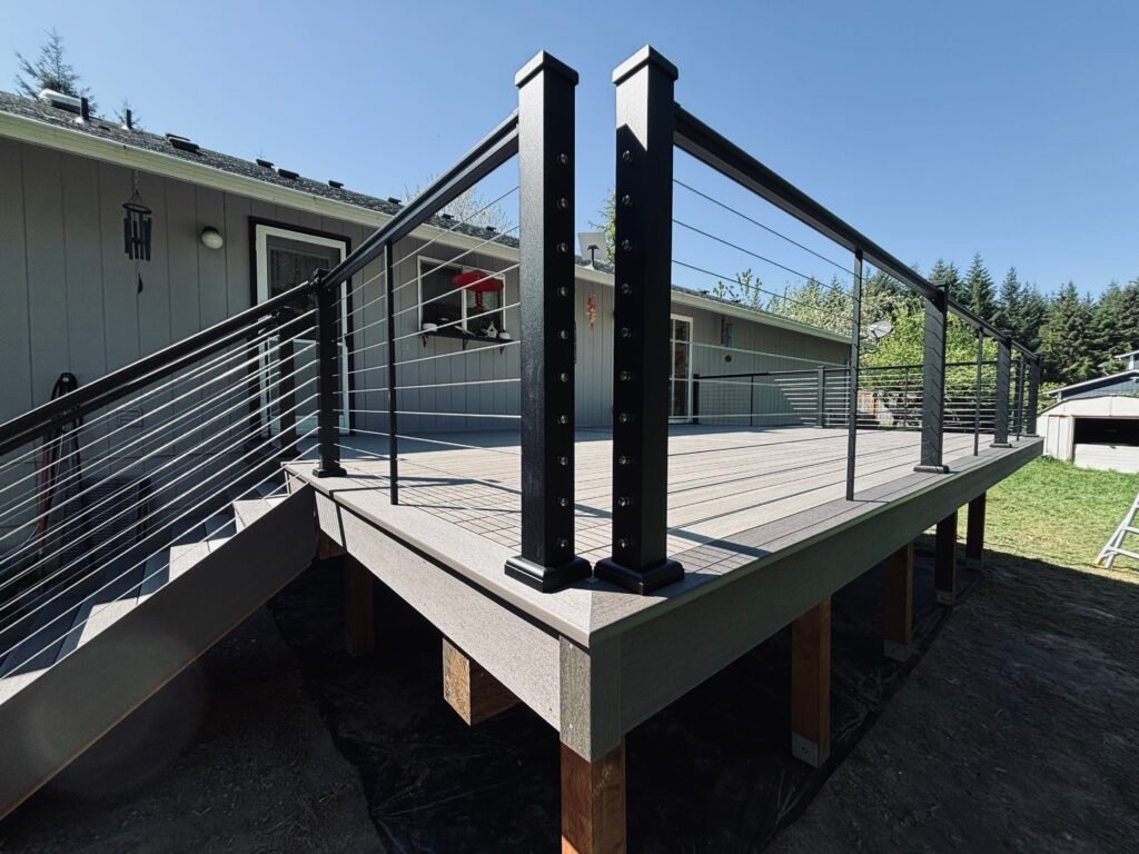 Modern cable railing on an elevated deck with black posts, aluminum rails, and a view of a gray house and trees under a clear blue sky.