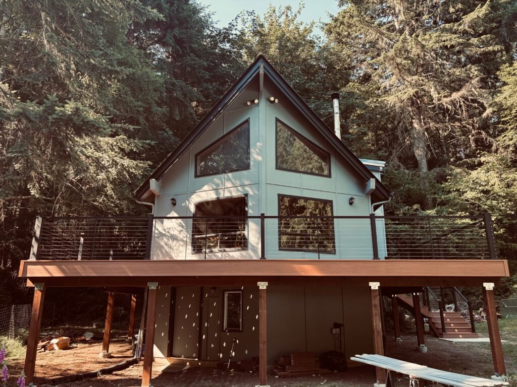 A modern A-frame cabin with large triangular windows, set among tall evergreen trees; wooden deck and metal railing in the foreground.